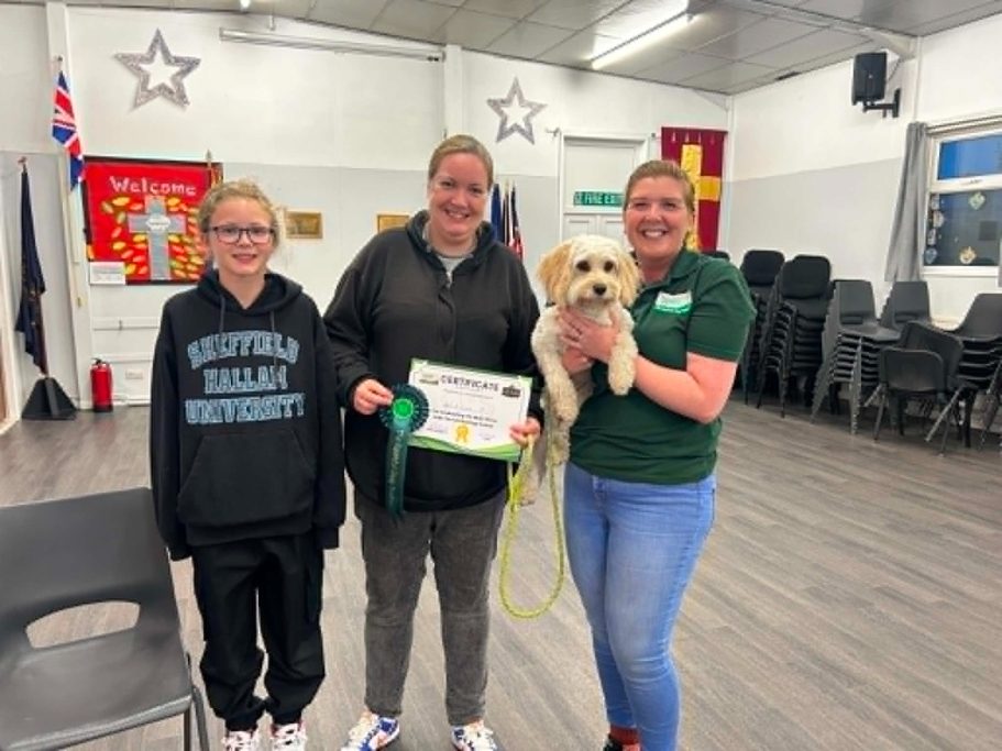 Three ladies in a dog training class. The trainer is holding the fluffy white dog and another lady is holding a certificate